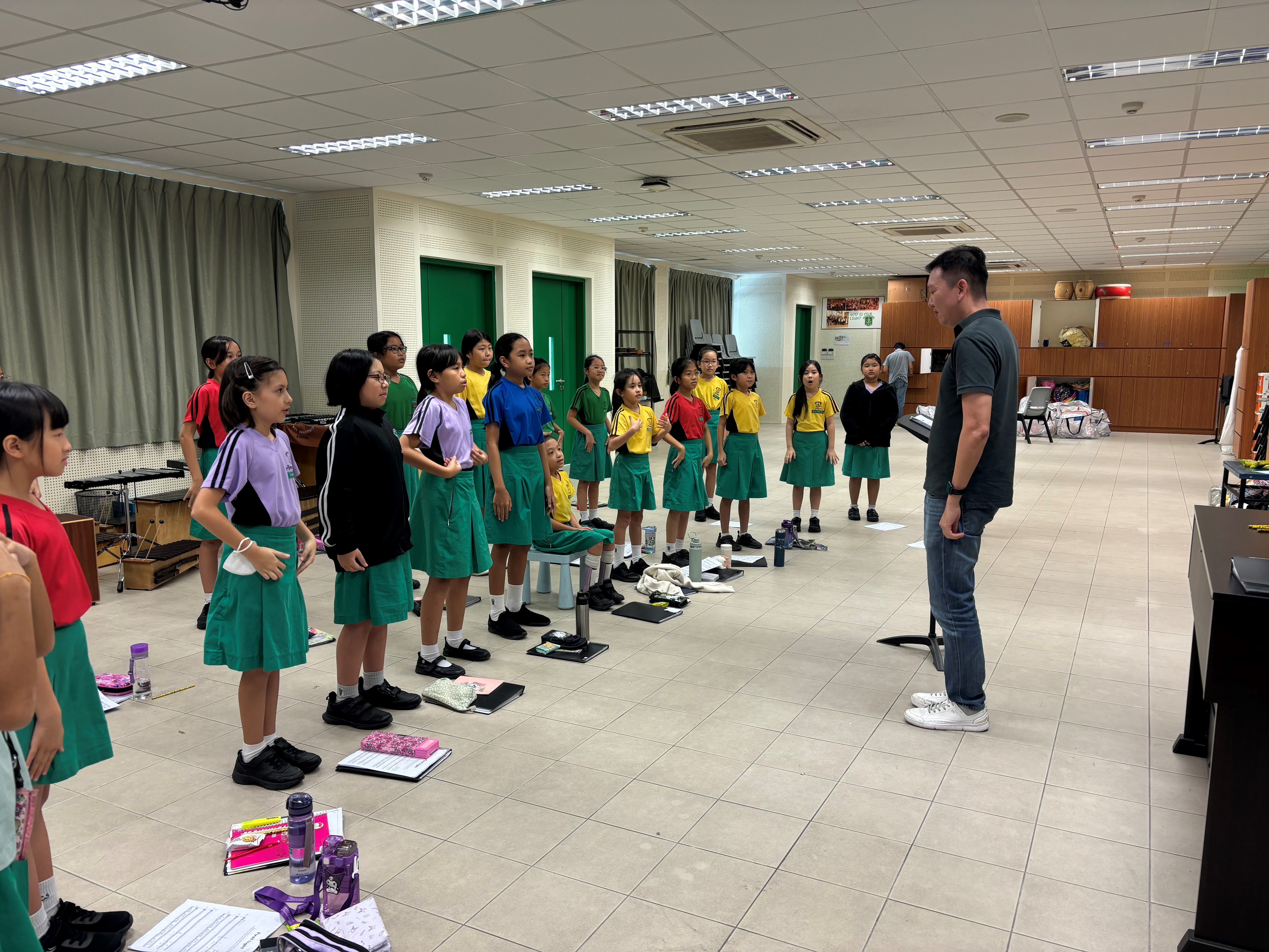 A man leads a choir rehearsal with young girls in green skirts standing in rows inside a school room.